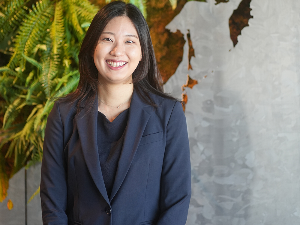 A businesswoman in a navy suit is smiling while standing in an office. The background features green plants and part of a decorative wall map.