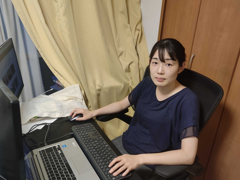 A woman is sitting at a desk working on a laptop and an external keyboard in an office or home office setting.
