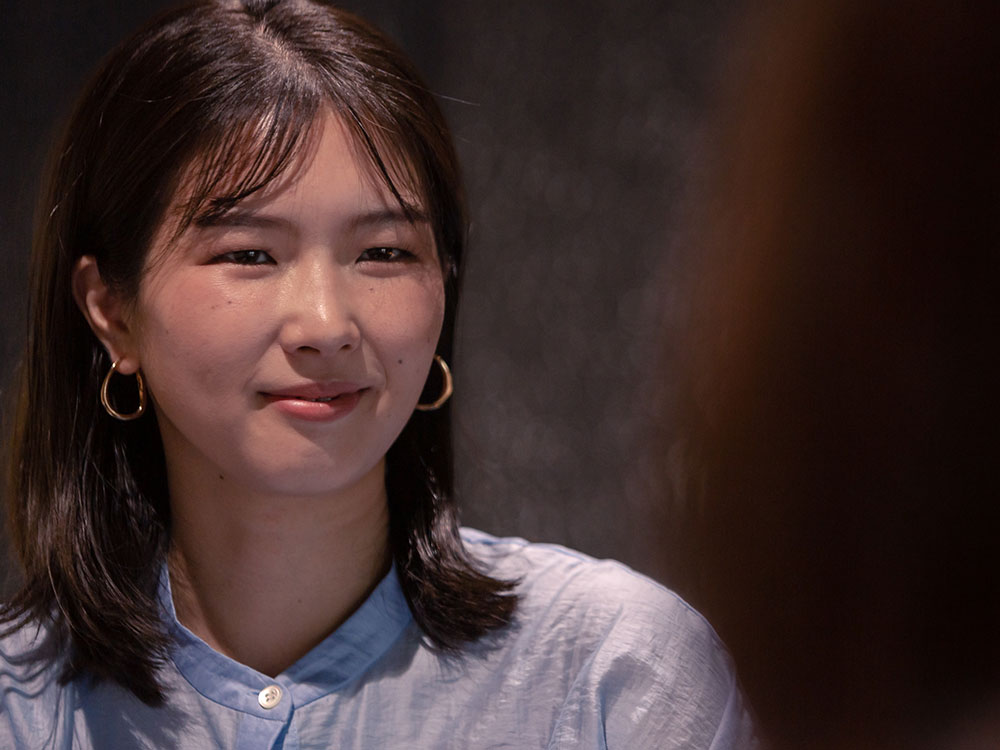 A woman smiling, wearing a light blue shirt and gold hoop earrings, with shoulder-length hair.