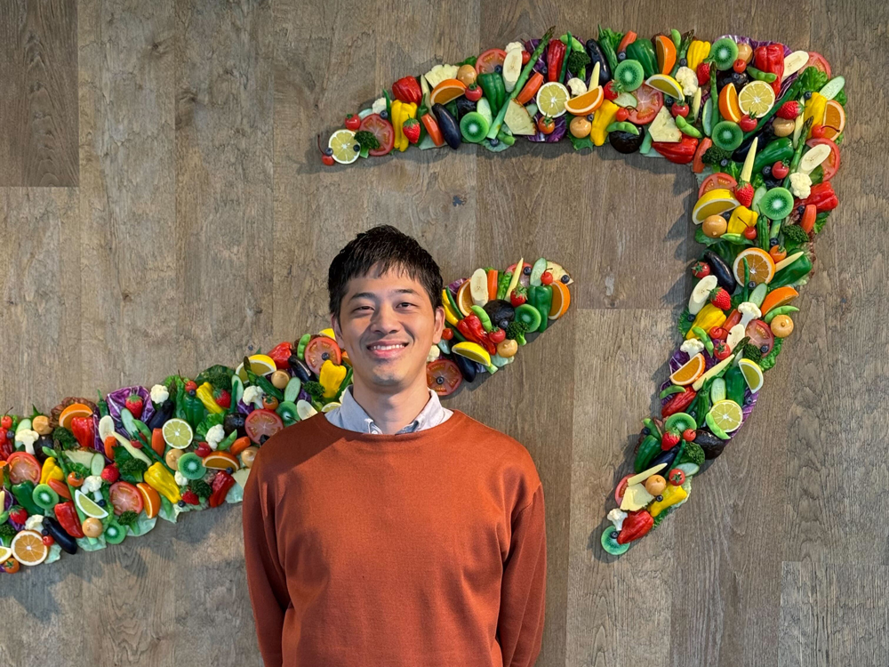 A man wearing an orange sweater stands in front of a wall decorated with colorful fruit and vegetable art.