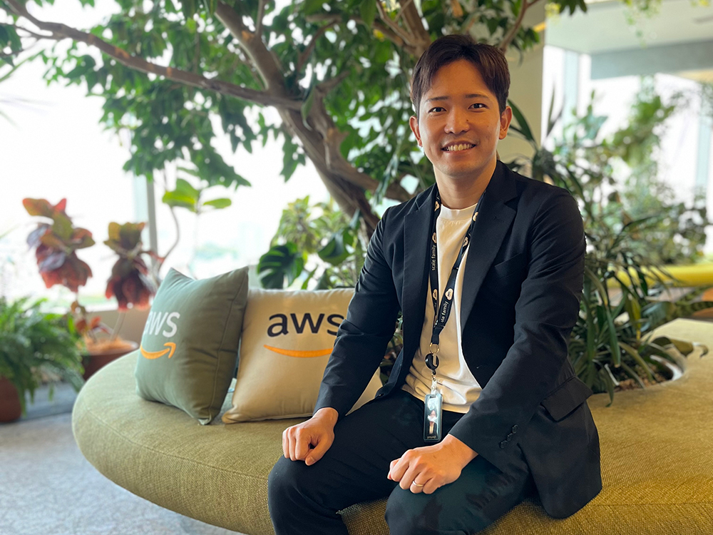 An AWS employee sitting on a green circular couch in a modern office environment with lush indoor plants and AWS-branded pillows visible in the background.
