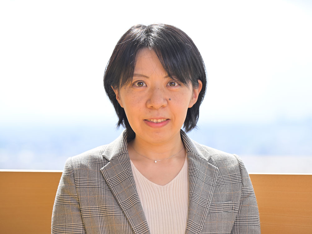 A woman with short black hair wearing a gray plaid blazer and light top, posing outdoors with a blurred background.