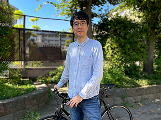A man standing outside next to a bicycle, with greenery and trees in the background.