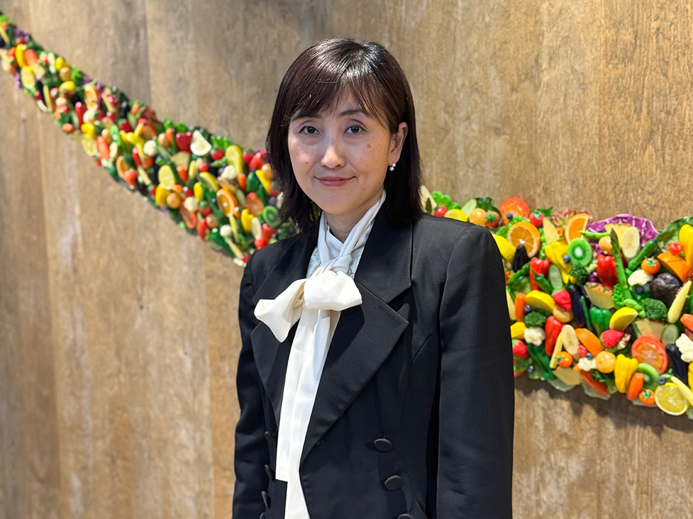 A woman in a black suit with a white bow blouse stands in front of an art installation made of colorful fruits and vegetables on a textured wall.