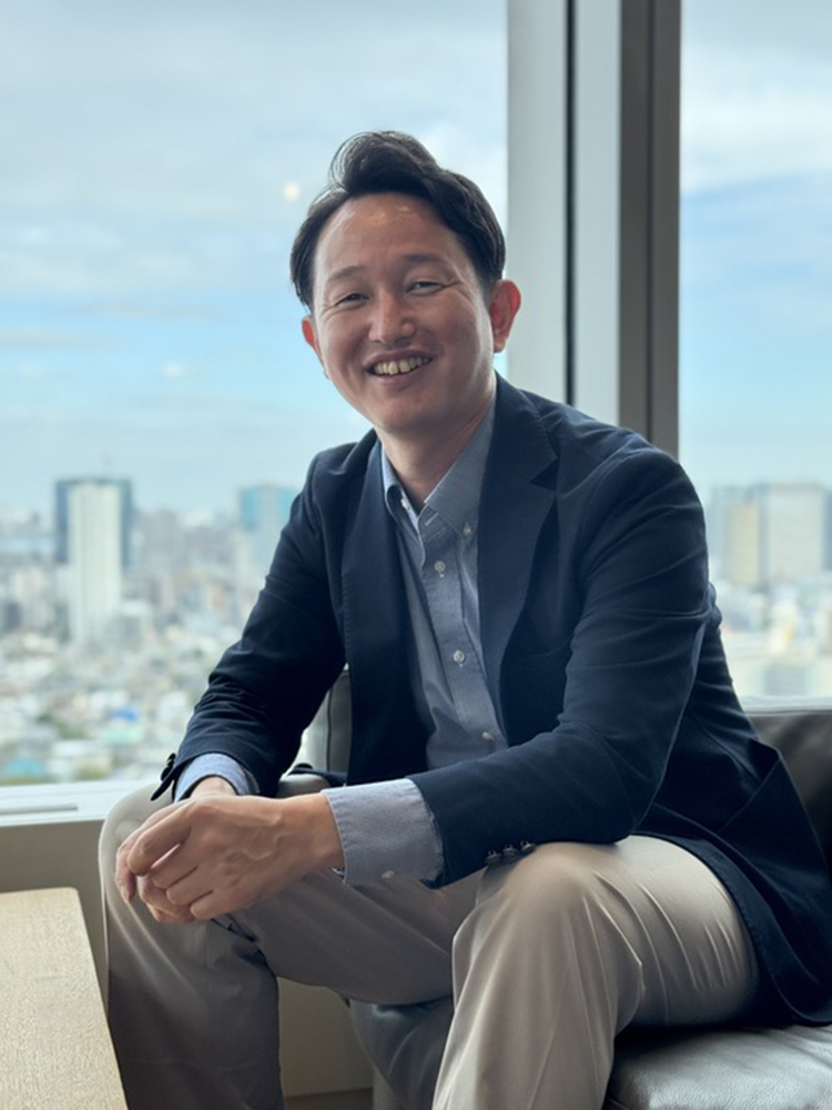 A smiling businessman sits by a window with a cityscape view in the background.