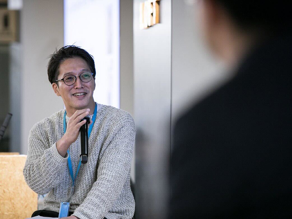A man wearing glasses and a light sweater is speaking into a microphone during a panel discussion. Another person is blurred in the foreground, suggesting an interactive or conversational setting.
