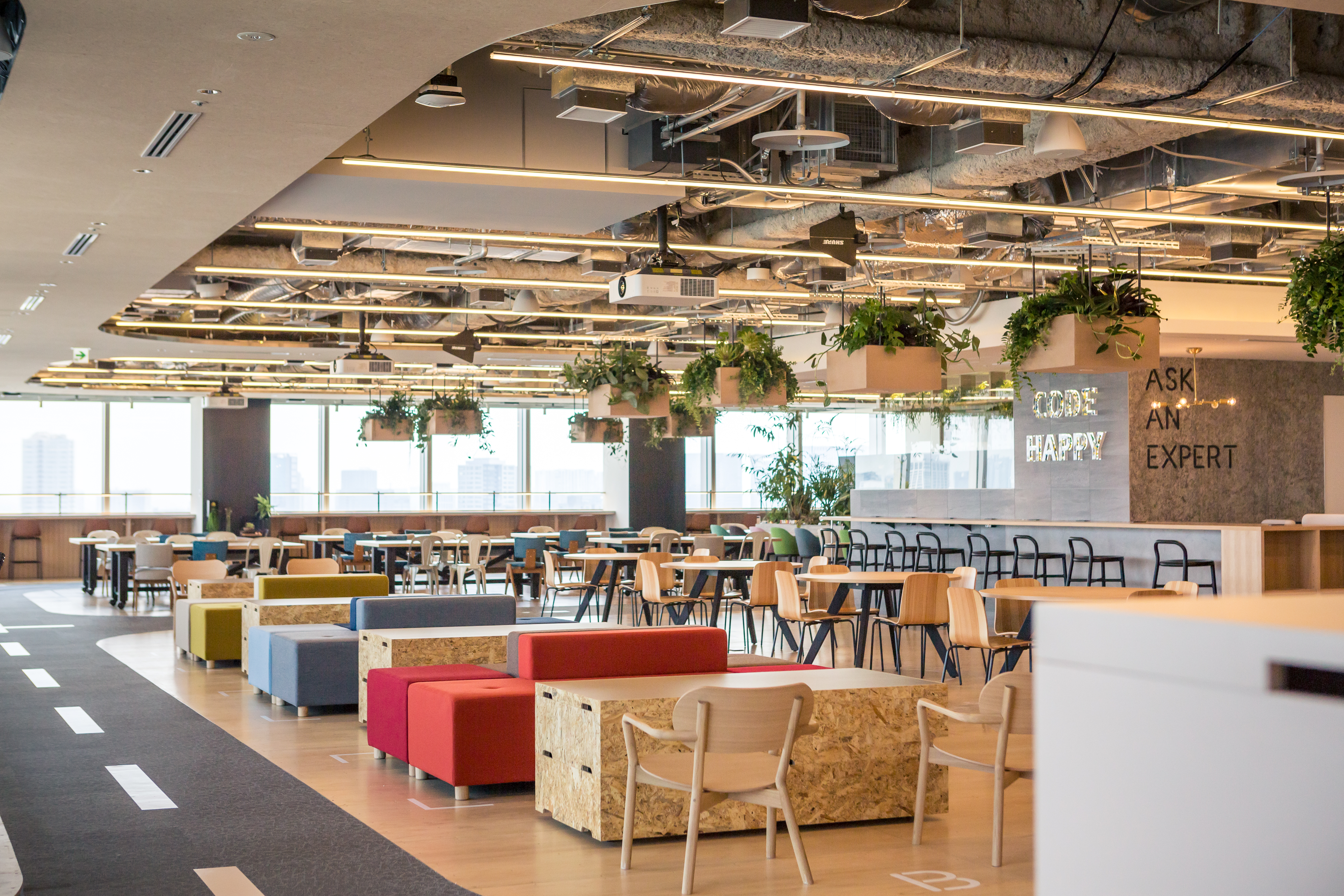 A modern open office environment featuring collaborative seating areas, natural light, overhead hanging plants, and signage reading 'CODE HAPPY' and 'ASK AN EXPERT' on the wall. The workspace has various colorful seating options and a relaxed, creative atmosphere.