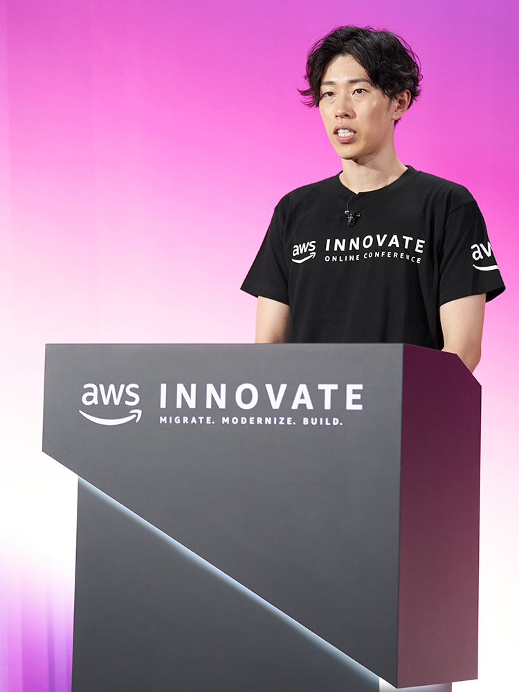 A speaker stands at a podium with the AWS Innovate Online Conference branding, set against a pink to purple gradient background. The speaker is wearing an AWS Innovate t-shirt and appears to be addressing an audience or participating in a virtual conference.
