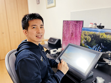 A person sitting at a desk in a home office, using a digital drawing tablet. Computer monitors and office equipment are visible in the background.