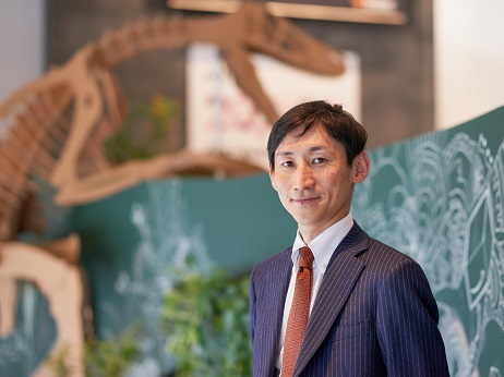 A business professional in a suit poses for a portrait with a dinosaur skeleton model and green chalkboard illustrations in the background.