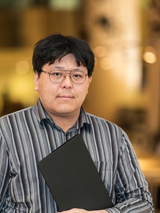 Portrait of an employee with glasses wearing a striped shirt and holding a black folder, posed in an office environment.