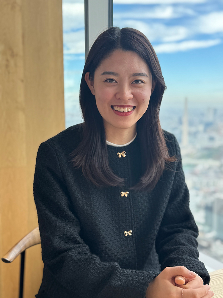 A woman is smiling while sitting by a window with a cityscape view in the background.