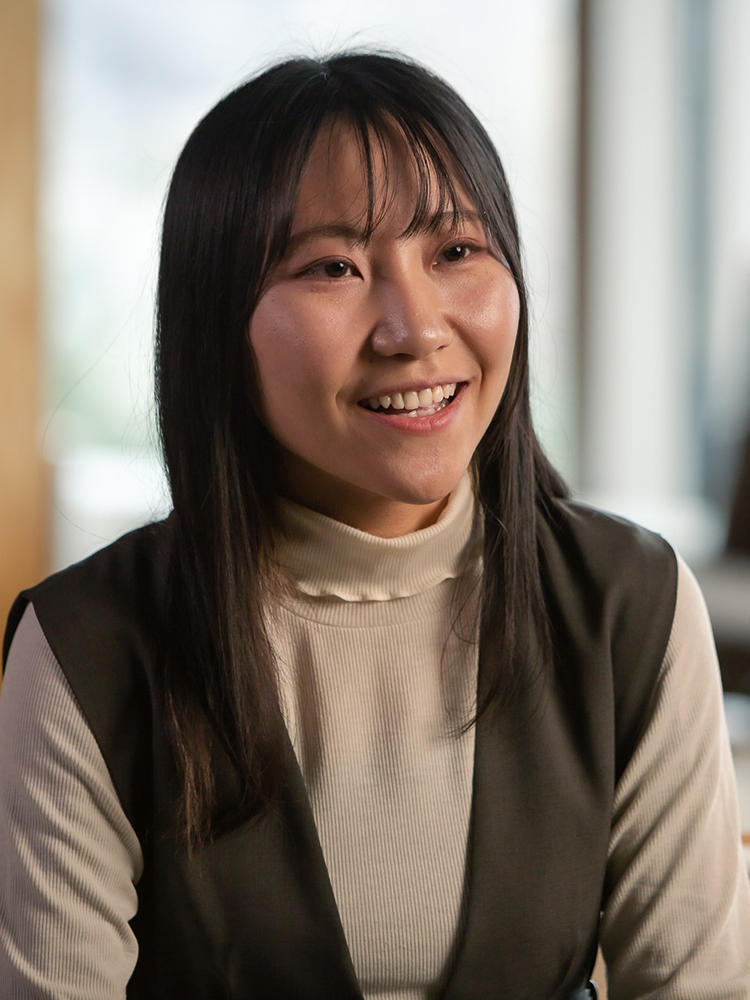 Portrait of a woman smiling indoors, wearing a light-colored turtleneck and dark vest.