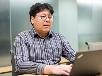 A person wearing glasses and a striped shirt is working on a laptop at a table in an office environment.