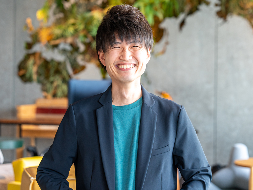 A man smiling broadly while wearing a casual blazer and green shirt, standing in a modern office with a creative, plant-filled background.