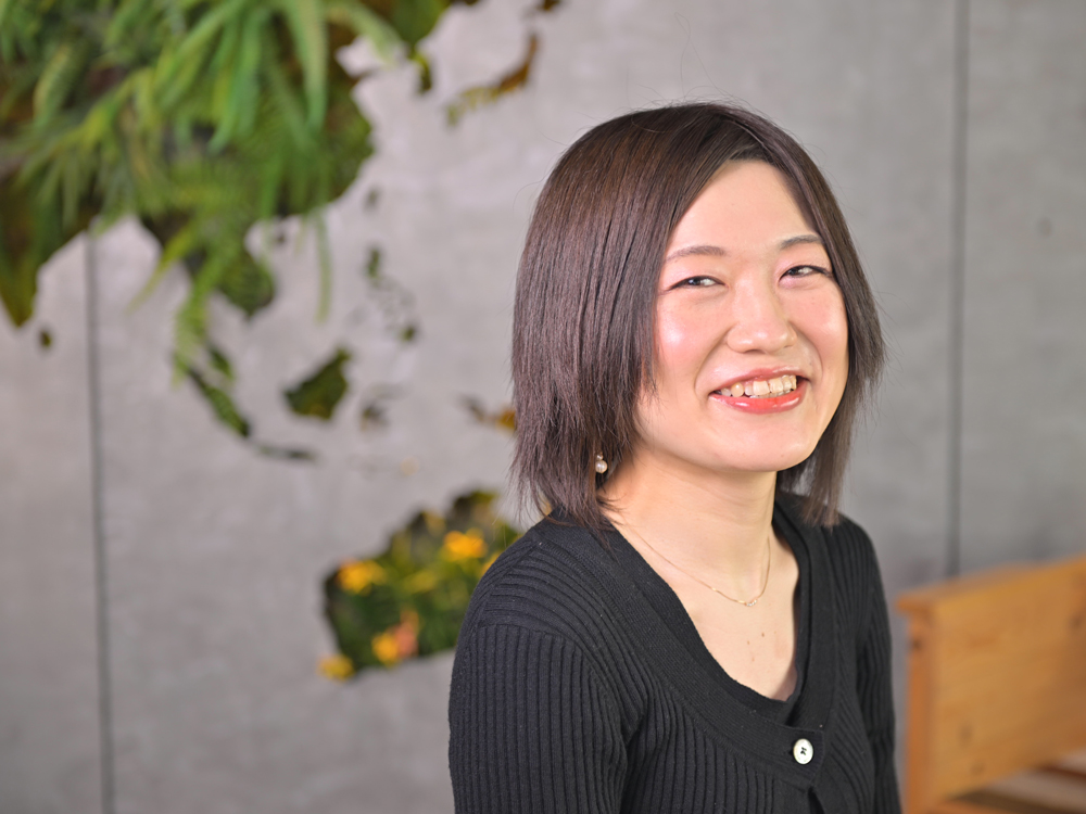 A woman with short hair is smiling indoors in front of a green wall background with plants.