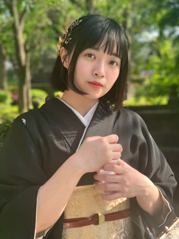 A woman in a traditional Japanese kimono standing outdoors, with greenery and sunlight in the background.
