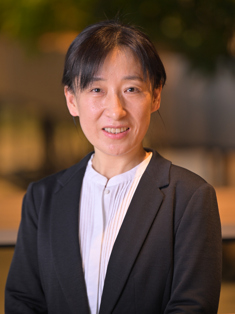 A professional portrait of a woman wearing business attire, featuring a black blazer and white blouse, set against a blurred office background.
