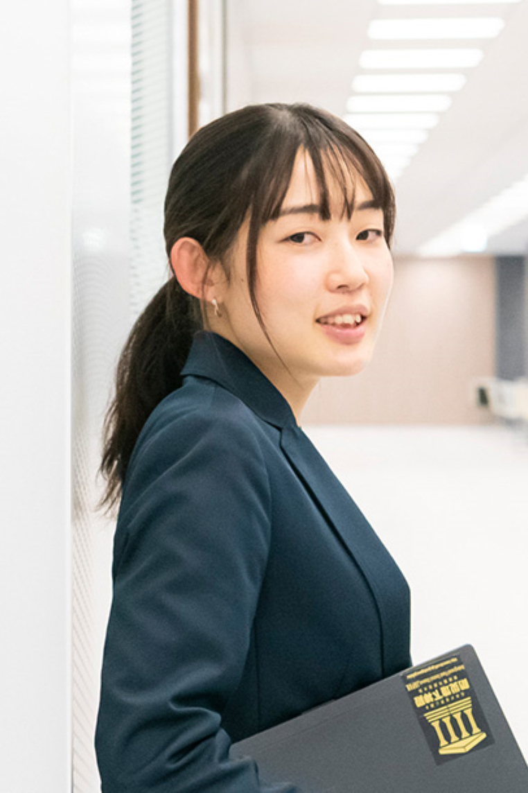 A businesswoman in a dark suit holding a folder, standing in a modern, bright office environment.