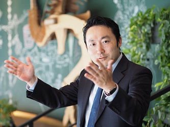 A business professional in a suit is gesturing with both hands while standing in a modern office environment with green plants and a decorative wall in the background.