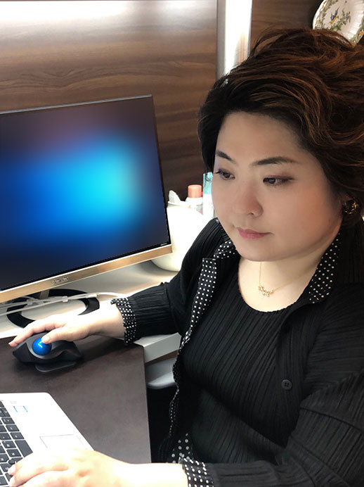 A woman is working at a desk with a computer, using a mouse and typing on a keyboard. She is focused on her work in an office environment.