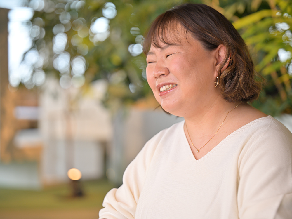 A portrait of a smiling woman wearing casual clothing, photographed outdoors with greenery in the background.