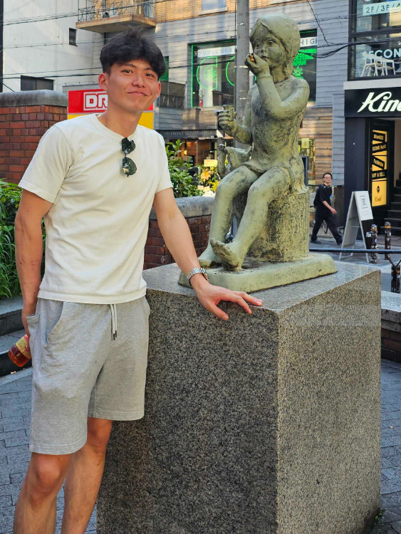 A man in casual clothing stands next to a statue of a child on a stone pedestal in an urban street setting.