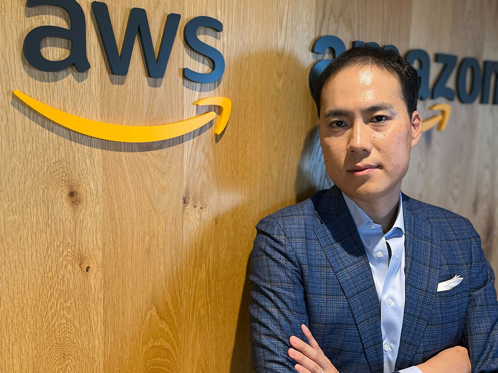 A businessman in a blue suit poses in front of a wooden wall with the AWS and Amazon logos.