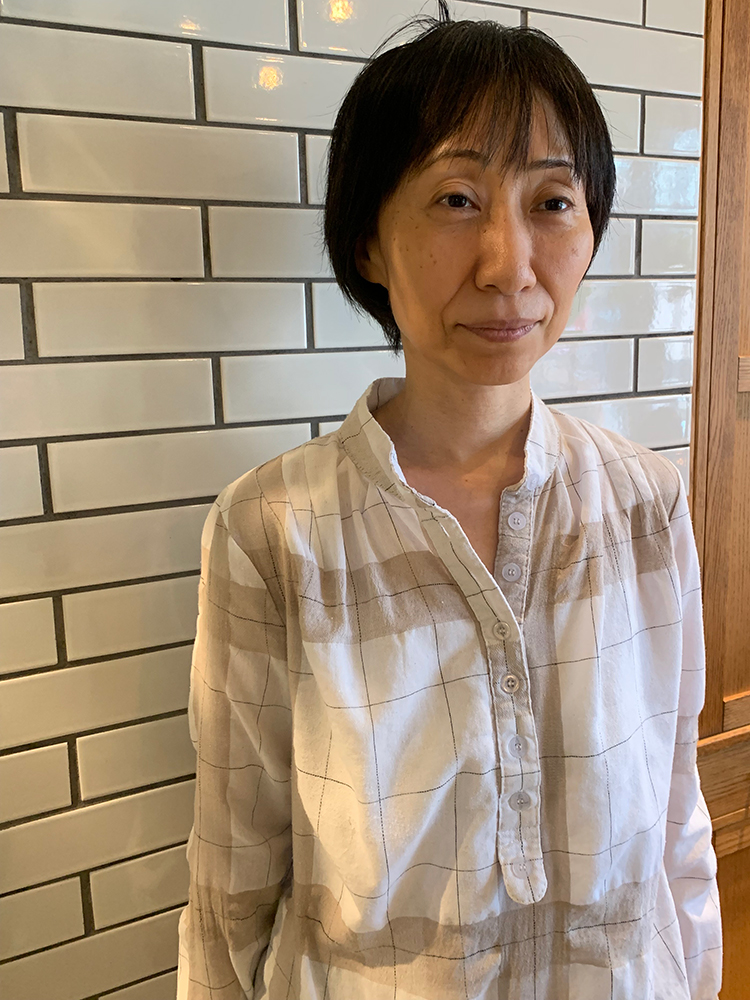 A woman stands in front of a white tiled wall, wearing a light-colored, checkered casual shirt. The setting appears to be indoors, and the atmosphere is casual.