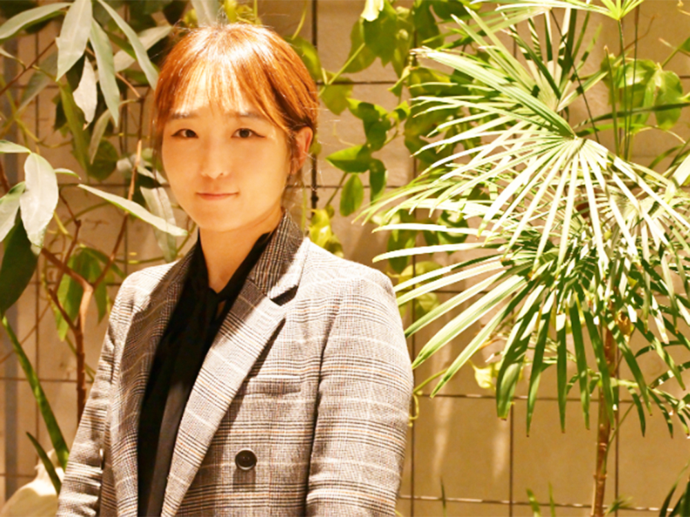 A woman wearing a plaid blazer standing indoors with various green plants in the background.