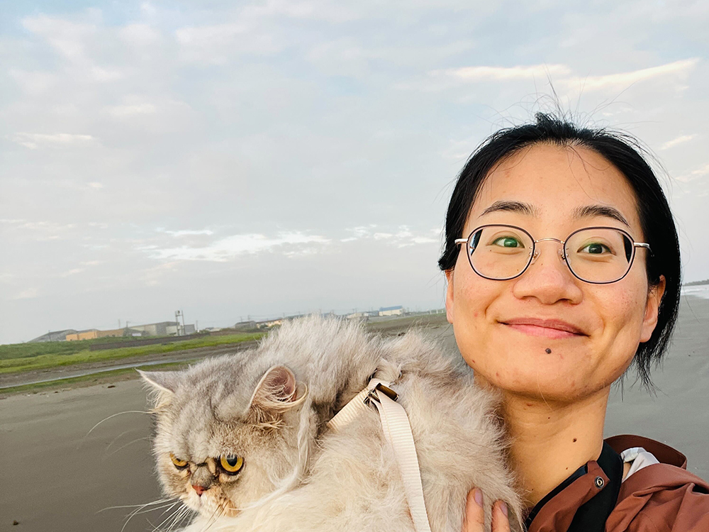 A woman with glasses holding a fluffy cat on a beach with overcast skies and distant buildings in the background.