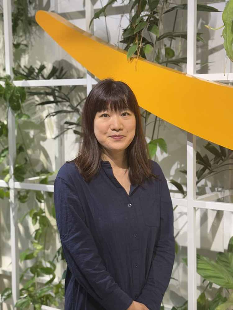 A woman stands in front of a modern indoor plant wall with a large yellow art piece in the background. She is wearing a dark shirt and smiling, with greenery and contemporary decor surrounding her.
