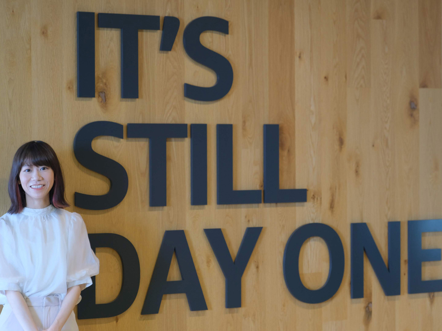 A woman in a white dress stands smiling in front of a wooden wall with large black letters spelling 'IT'S STILL DAY ONE'.