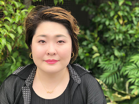 Portrait of a woman with short hair, wearing a black top and gold earrings, outdoors in front of lush green foliage.