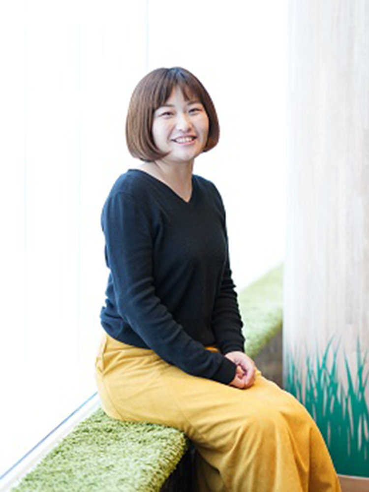 A woman in a black top and yellow skirt is sitting by a window on a green cushioned ledge, smiling at the camera.