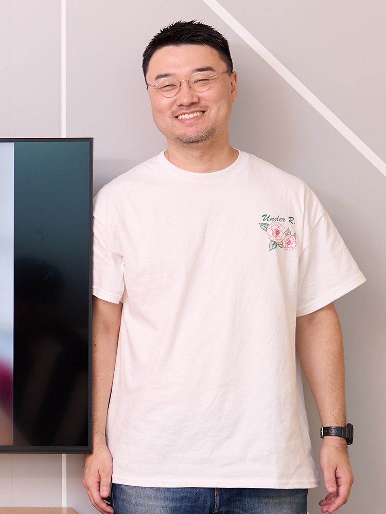A smiling man wearing a white t-shirt with a floral design and glasses, standing casually indoors.