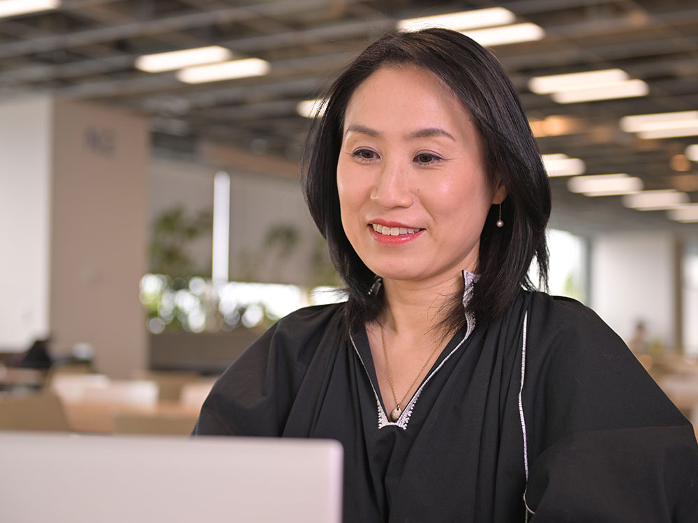 A woman smiling while working on her laptop in a modern, open office environment.