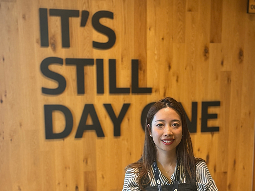 A woman stands smiling in front of a wood-paneled wall with a large sign that reads 'IT'S STILL DAY ONE.'
