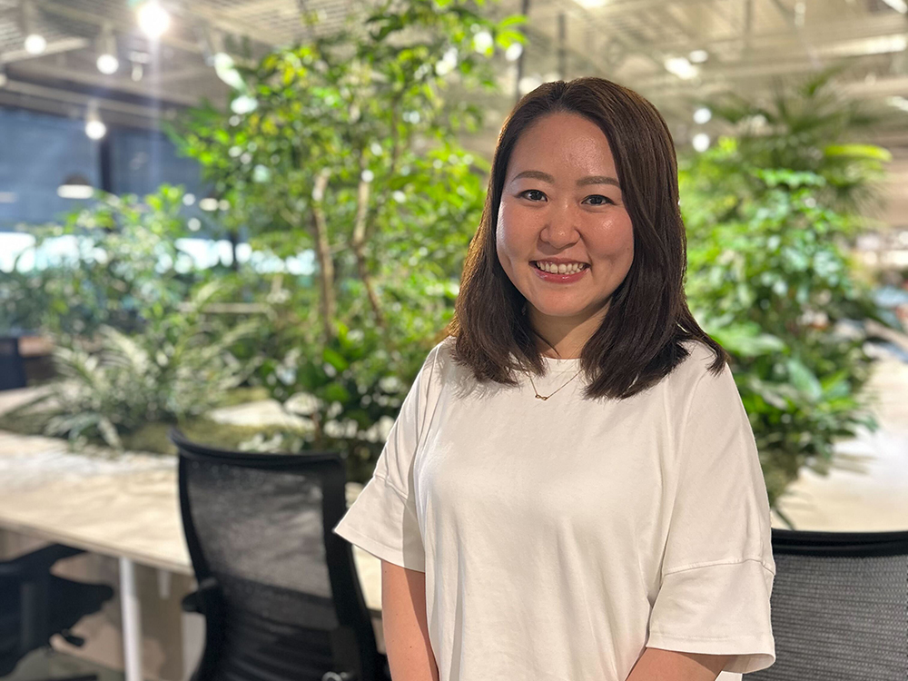 A woman smiling in a modern office environment with abundant greenery in the background.