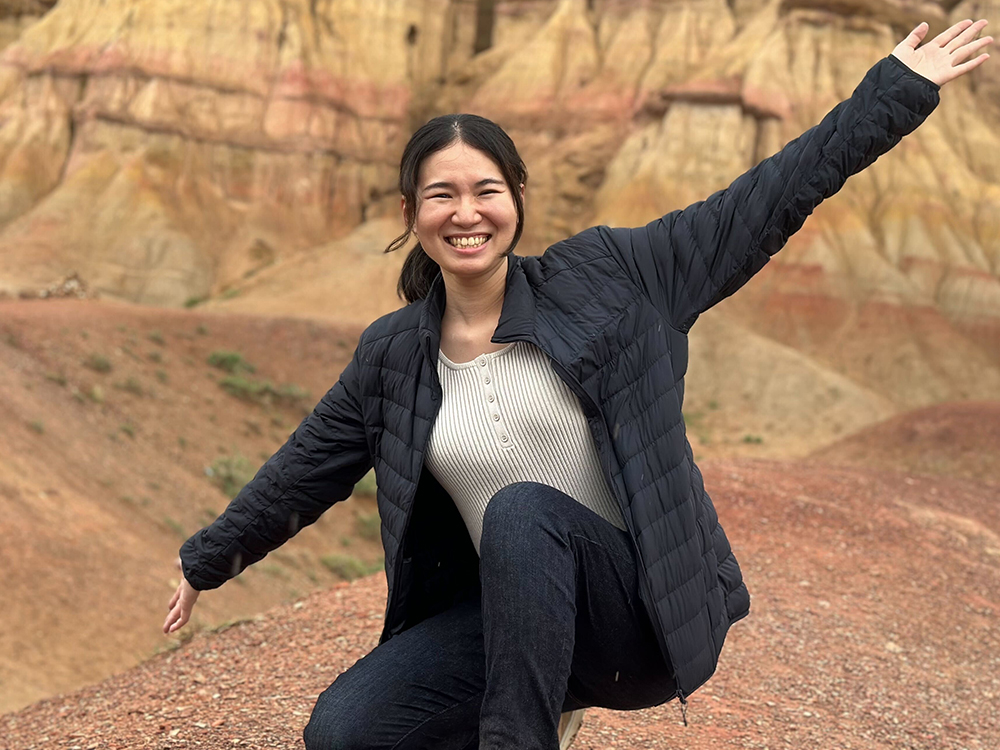 A smiling woman with arms outstretched poses in front of a background of colorful rock formations.