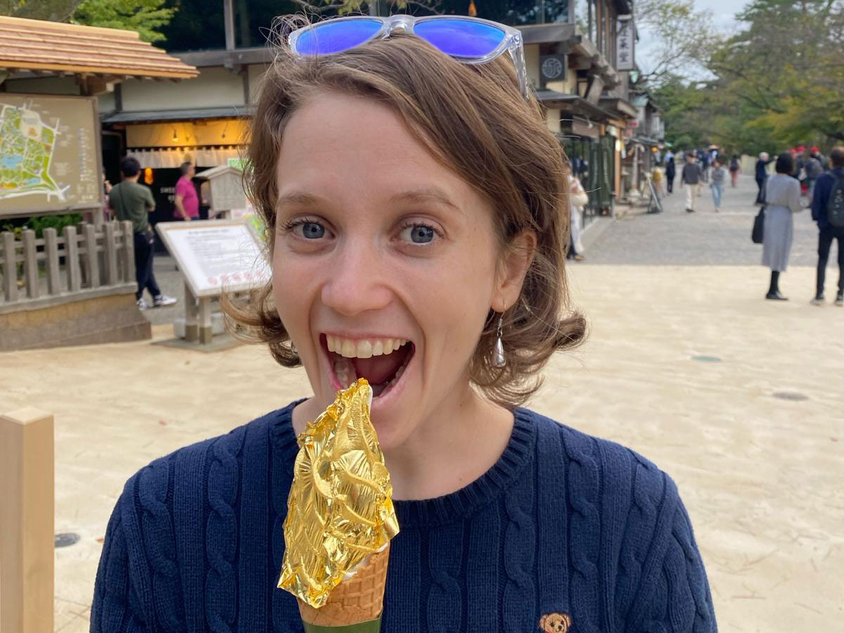 A woman enjoying a gold leaf-covered ice cream cone outdoors, with a lively outdoor street scene in the background.