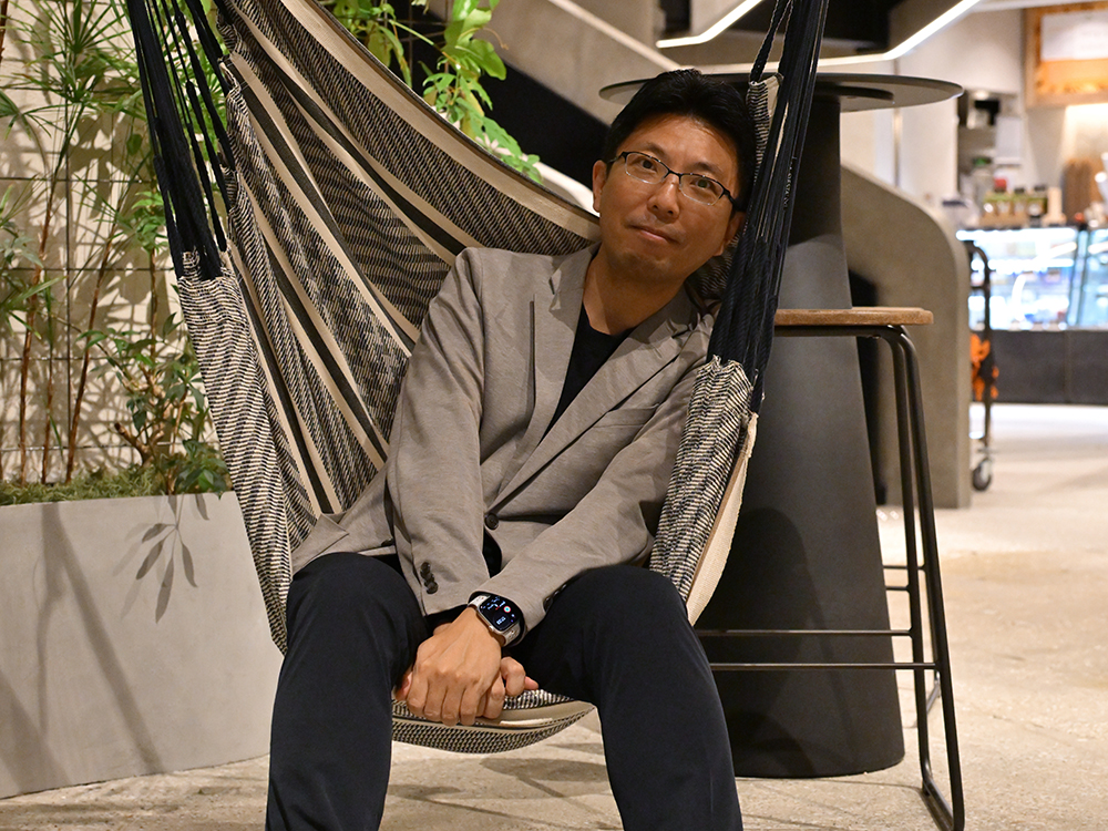 A man wearing glasses and a gray blazer is sitting in a hammock inside a modern cafe. The interior features contemporary decor with plants and stylish furniture.