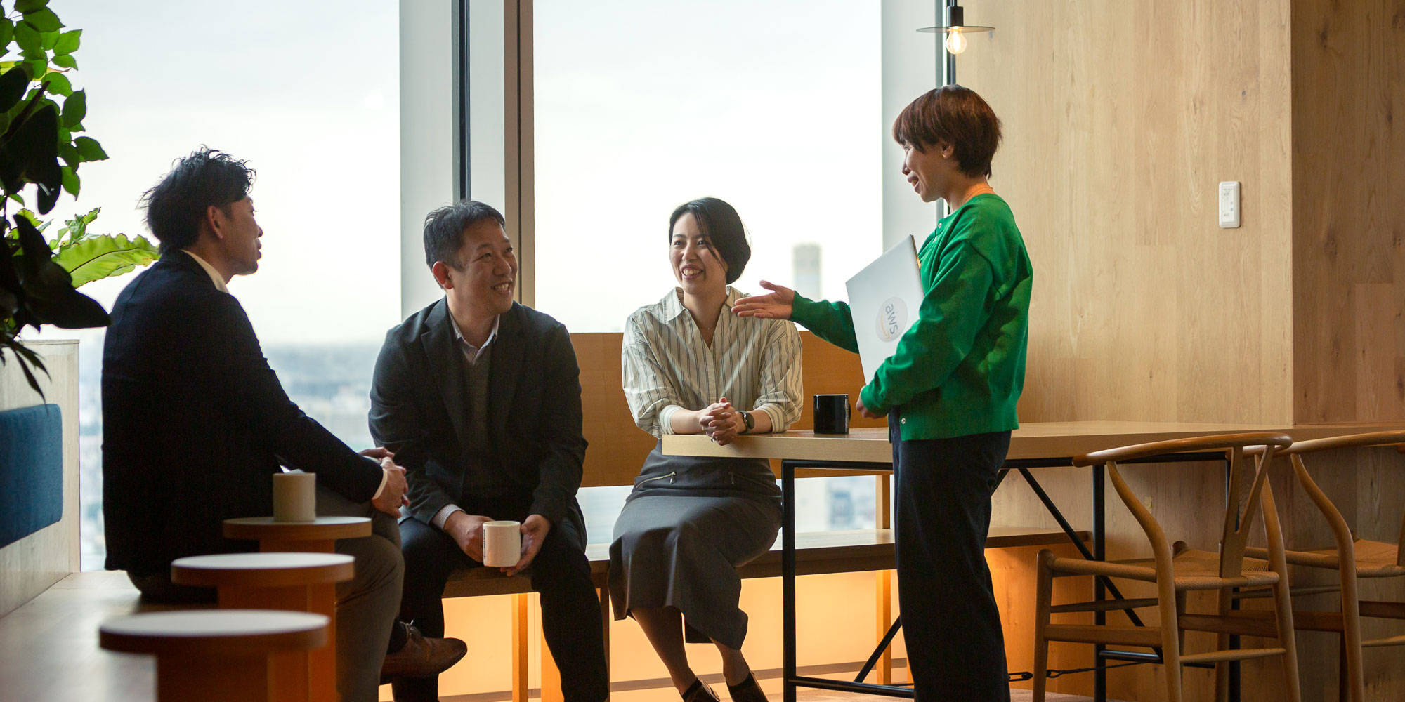 Four people having a business meeting in a modern office setting, with large windows and city views. The group appears engaged in a casual and collaborative discussion.