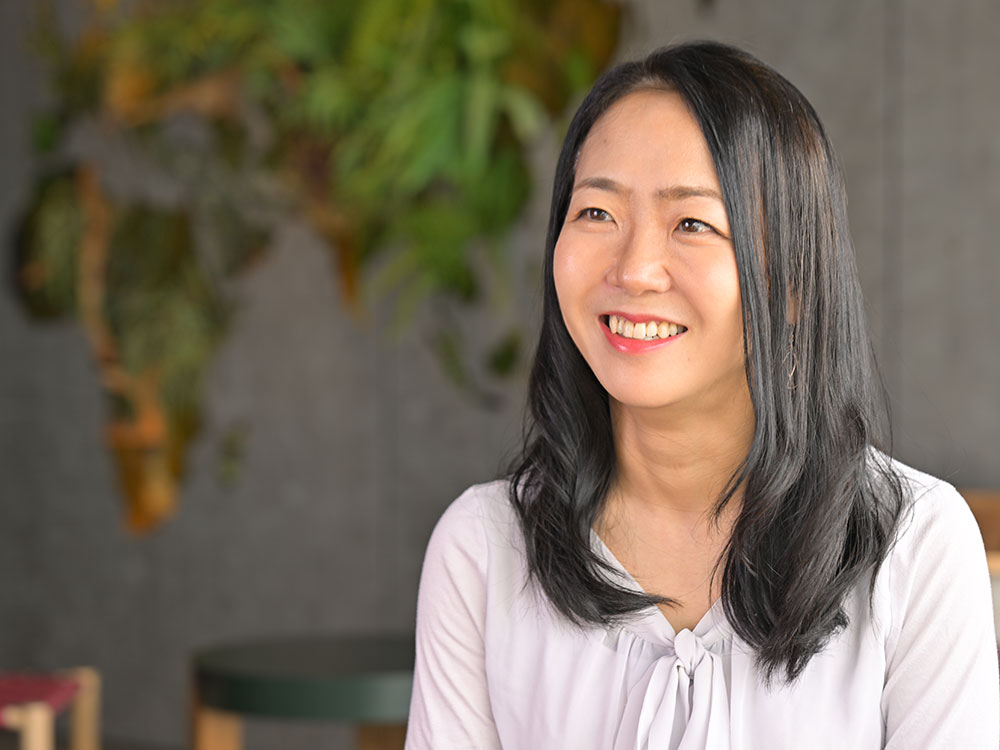 A cheerful woman with long dark hair is smiling indoors, wearing a light-colored blouse, with a blurred abstract background.
