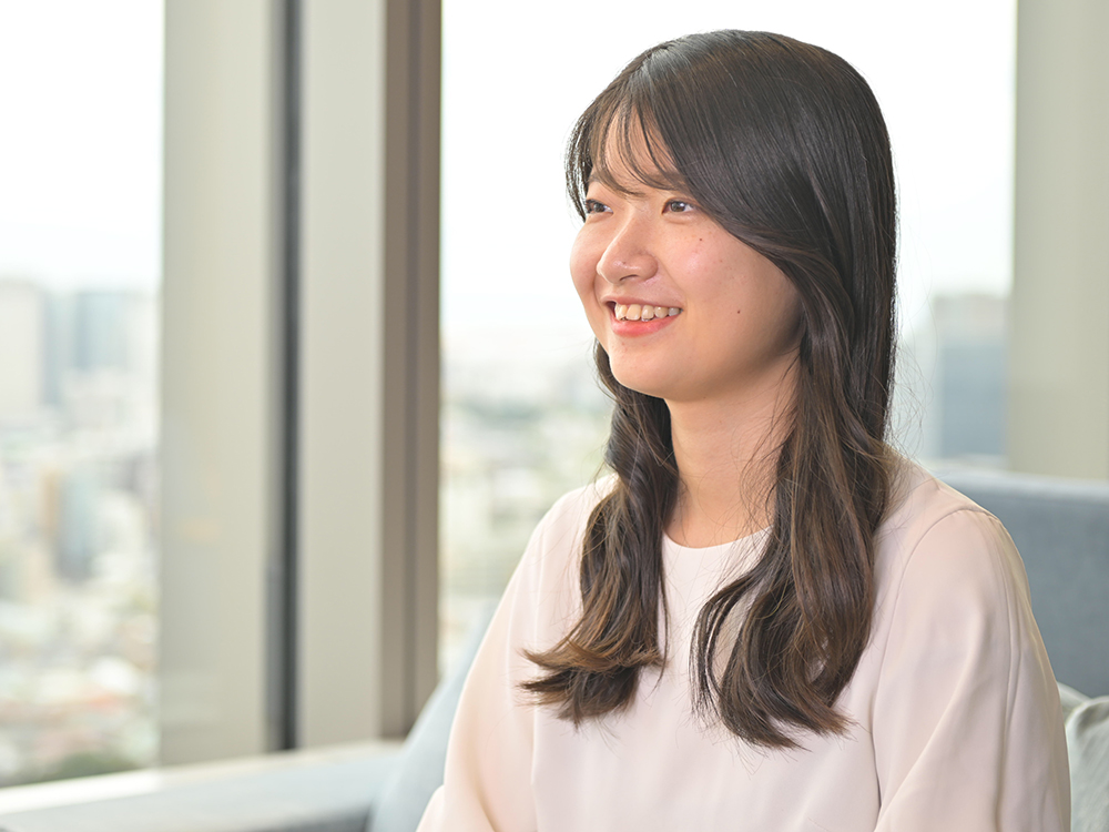 A young woman with long hair is smiling while sitting indoors by a large office window, with natural light illuminating the scene.