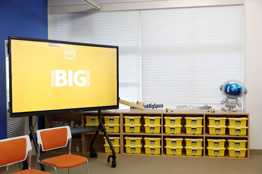 A classroom setup at AWS Think Big Space featuring a large screen displaying the 'Think Big Space' logo, yellow storage bins labeled 'BIG,' orange chairs, and a small educational robot with a blue face screen. The environment is bright and designed for learning and community engagement.