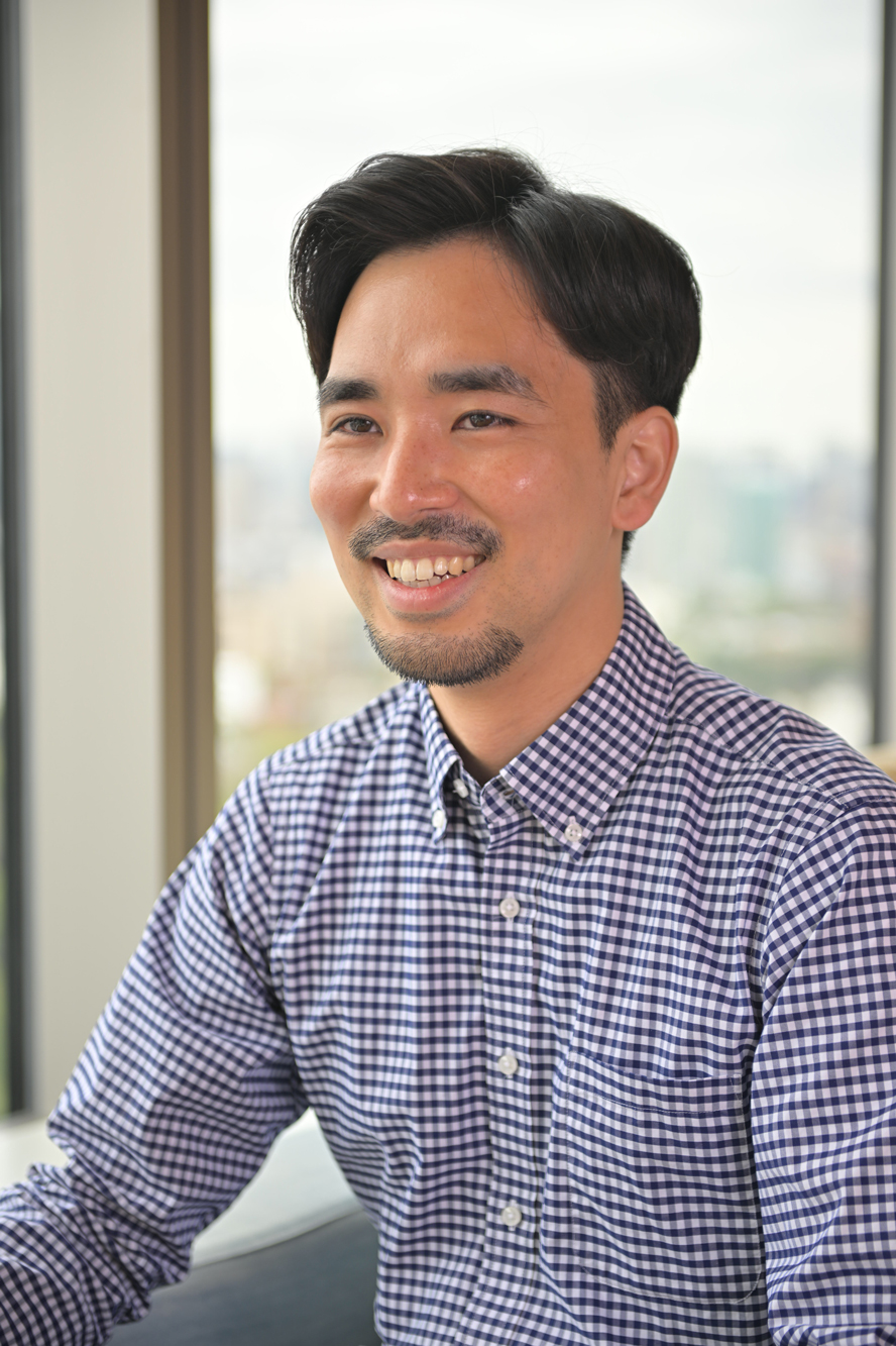 A smiling man wearing a checkered shirt, seated in an office setting with large windows and a city view in the background.