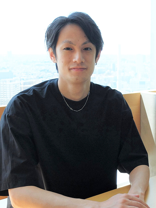 A young man wearing a black shirt sits by a window with a city view in the background. He has short dark hair and is facing the camera, looking relaxed and casual.
