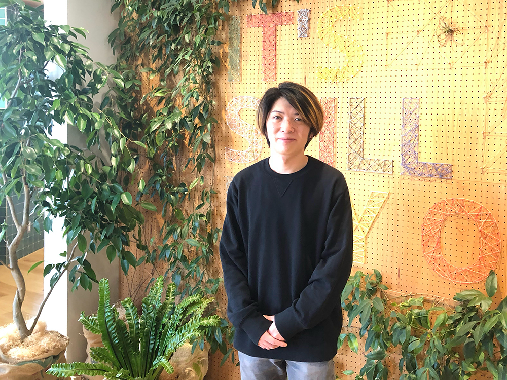 A person stands in front of a wall decorated with string art and surrounded by lush indoor plants. The background features colorful letters created with string art.