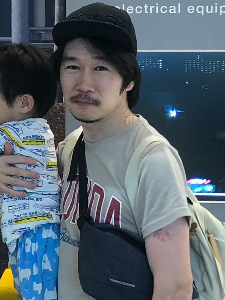 A man wearing a casual outfit and a backpack is holding a child. Both are dressed casually and appear to be in a public indoor setting.